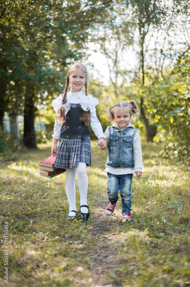 Fototapeta premium A schoolgirl walks in the park by the hand with her younger sister and carries books in her hand.