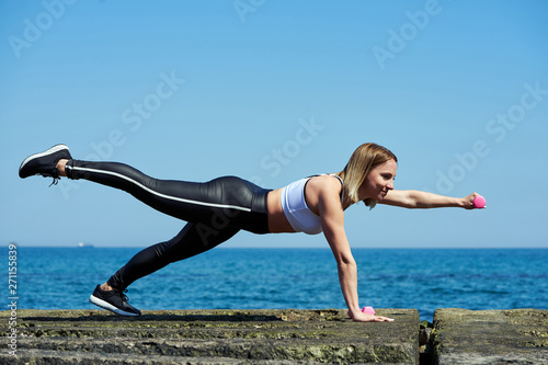 Horizontal photo of a beautiful woman who does sports exercises on the beach of the ocean or sea. The woman is holding dumbbells in her hands.