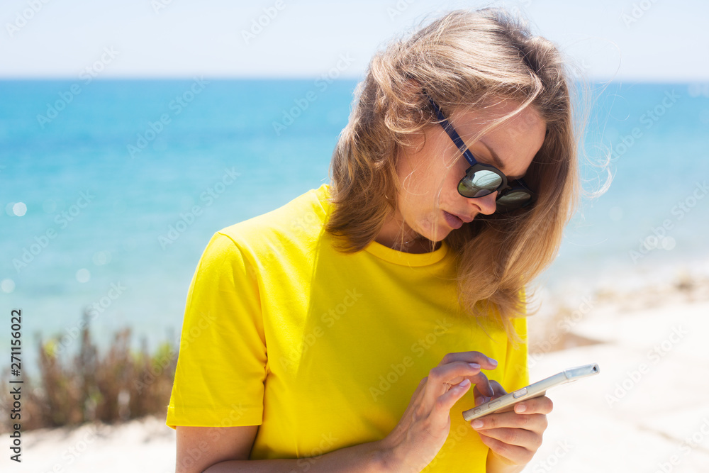 Beautiful woman wearing in t-shirt, blue jeans and sunglasses, staying near coast line. She uses her mobile phone