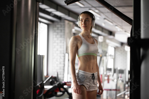 Closeup portrait of young pretty and muscular woman in gym listening the music in headphones. Stylish black interior of modern fitness gym. Concept of health and sport lifestyle. Athletic Body.