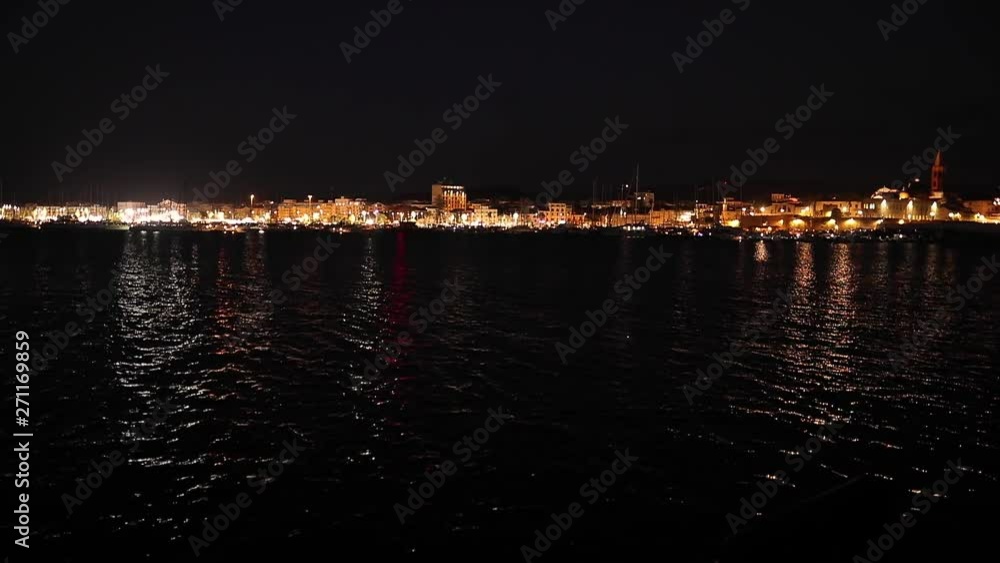 Illuminated night cityscape view across Alghero's coastline mirrored by rippling ocean waves below. Sardinia, Italy. Illuminated night cityscape view across Alghero's coastline mirrored by rippling ocean waves below. Sardinia, Italy.