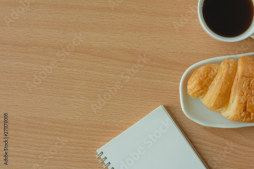 Top view of Croissant, cup of hot coffee and notebook with copy space on wood background.