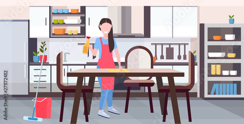 housewife in apron wiping dinning table by dust cloth girl doing housework cleaning service housekeeping concept full length flat modern kitchen interior horizontal
