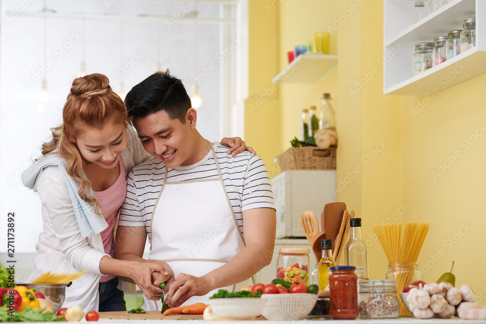 Smiling young Asian woman helping husban with cutting vegetable for salad or soup
