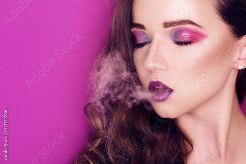 Fashion model woman creative colorful pink and blue make up. Beauty portrait of beautiful girl. Smoking girl, Close up of a female inhaling from an electronic cigarette. Purple background