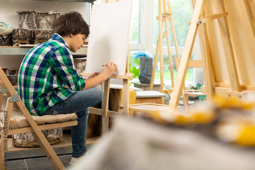 Dark-haired boy sitting near painting easel and drawing