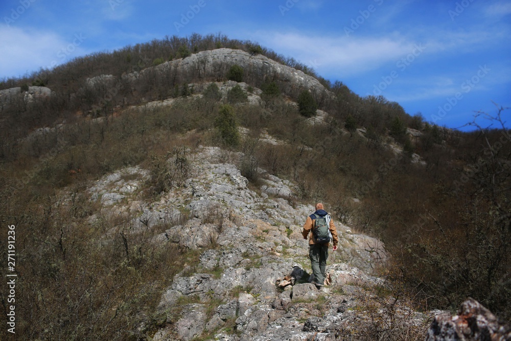 Redhead man in a brown-and-blue windbreaker with backpack walks on a rocky hillside in the spring Crimean forest. There are no leaves on the trees yet. Travel, adventure and hiking concept.