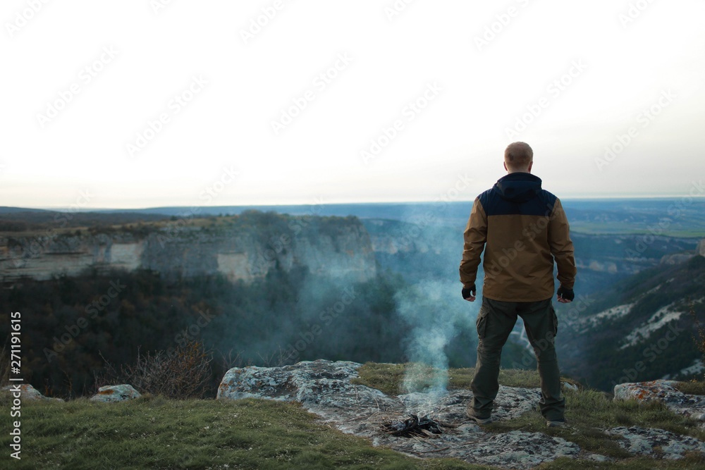 Redhead man in a brown-and-blue windbreaker stands back to camera near an extinct fire, from which there is still smoke, on the Mangup plateau in the Crimea. Travel, adventure and hiking concept.