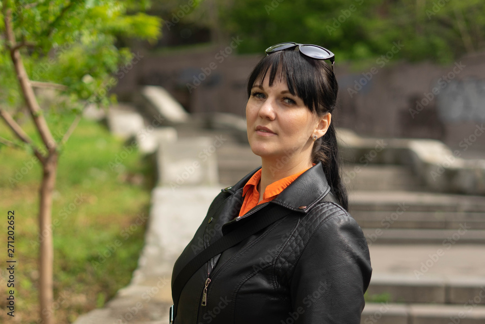 A beautiful brunette woman in a black jacket and an orange shirt is posing against the background of a long concrete staircase in the park. A beautiful brunette woman in a black jacket and an orange shirt is posing against the background of a long concrete staircase in the park.
