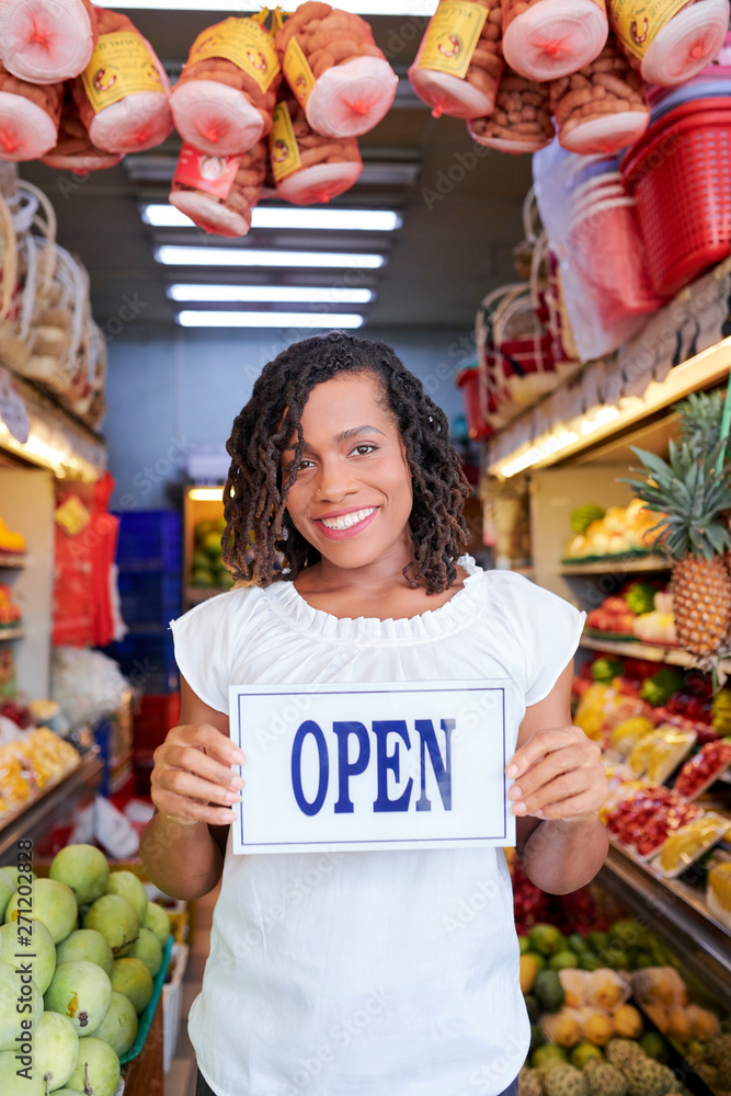 Portrait of happy smiling young woman standing in fruit and vegetable shop with open signboard in hands