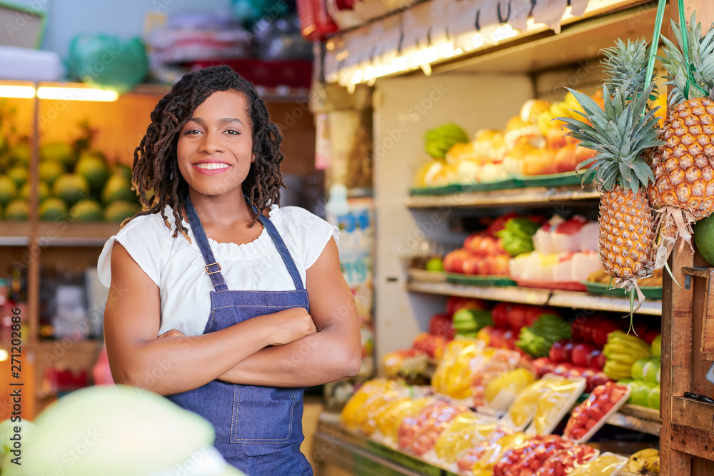Portrait of pretty smiling Black saleswoman standing in grocery store with her arms crossed