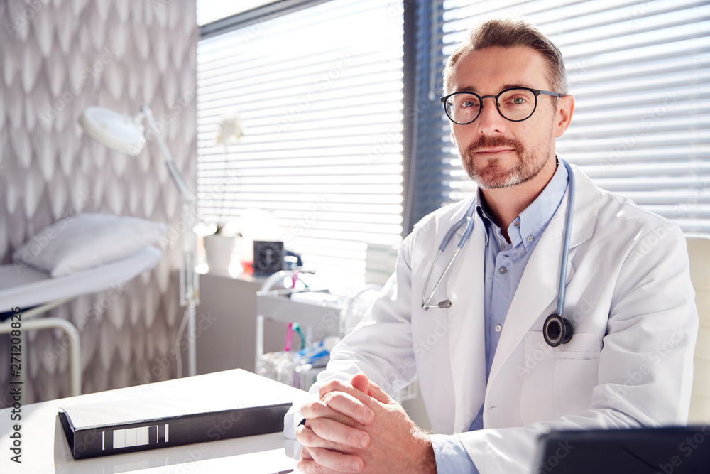Portrait Of Smiling Male Doctor Wearing White Coat With Stethoscope Sitting Behind Desk In Office Portrait Of Smiling Male Doctor Wearing White Coat With Stethoscope Sitting Behind Desk In Office