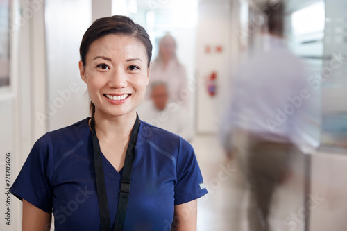 Portrait Of Smiling Female Nurse Wearing Scrubs In Busy Hospital Corridor