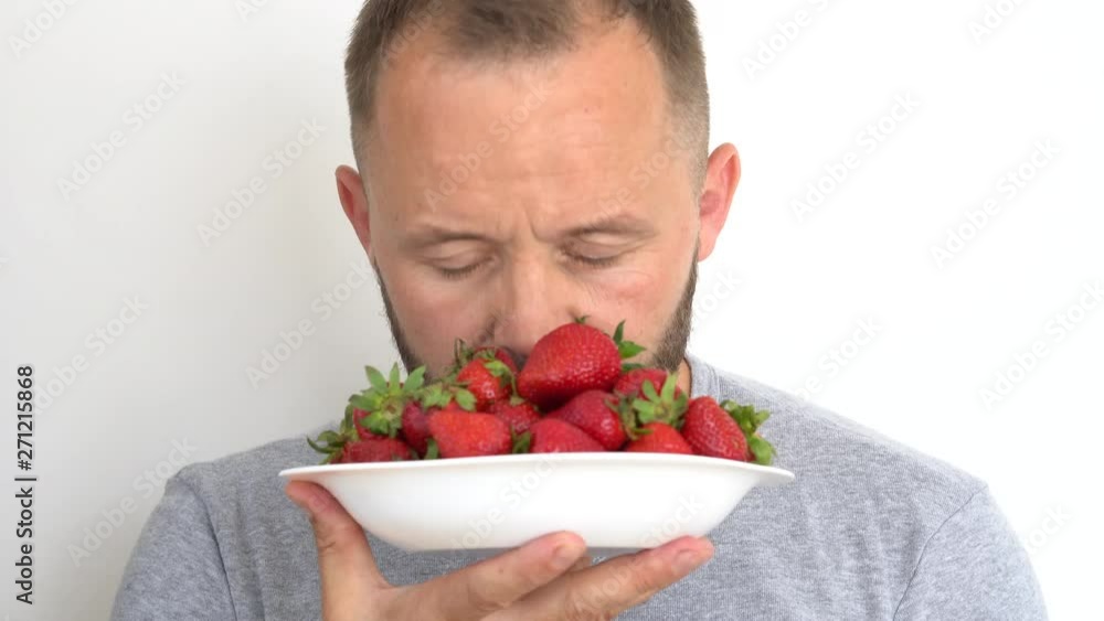 close-up of a young man with a short haircut holding a white plate full of ripe bright red strawberries with pleasure inhaling the scent of her close-up of a young man with a short haircut holding a white plate full of ripe bright red strawberries with pleasure inhaling the scent of her