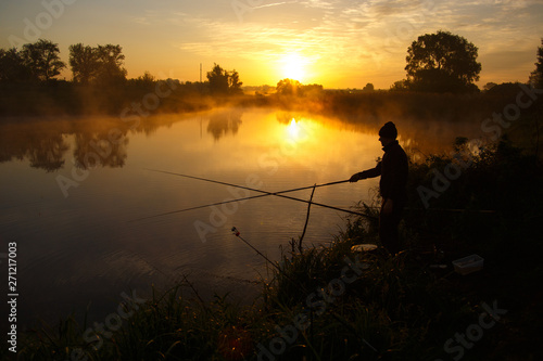 Lonely fisherman angling at foggy lake in the early morning just after golden sunrise