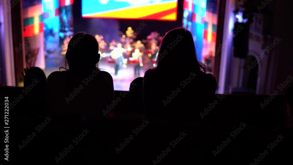 Silhouettes of two women who sit in the auditorium and watch a performance on stage. Silhouettes of two women who sit in the auditorium and watch a performance on stage.