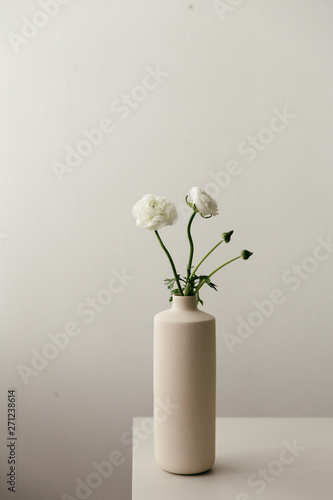 White ranunculus in a tall beige vase in a moody setting.