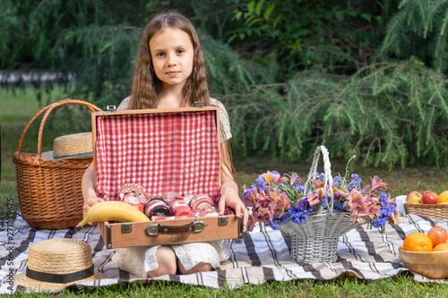 Portrait of a beautiful girl with a basket of flowers on a picnic in the park. The child holds open a chest with jam