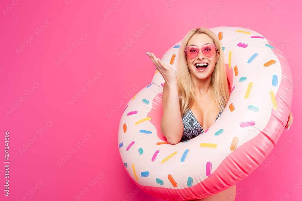 Image of girl in swimsuit sunglasses with inflatable donut for swimming on empty pink background. Indicates with hand to side Image of girl in swimsuit sunglasses with inflatable donut for swimming on empty pink background. Indicates with hand to side