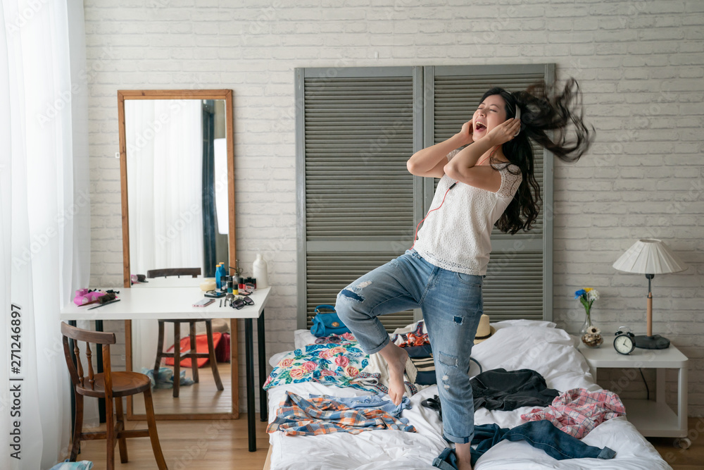 Excited woman listening to music and dancing in home bedroom. crazy funny girl wear headphones enjoy melody new songs on internet move body relax while packing prepared luggage with clothes on bed. - obrazy, fototapety, plakaty 