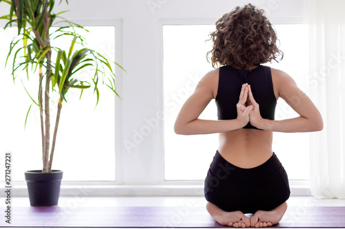 Back view of girl is sitting in vajrasana diamond pose with hand folded in namaste. Woman is practicing yoga, meditating in reverse prayer pose paschima namaskarasana at studio. Healthy life concept.