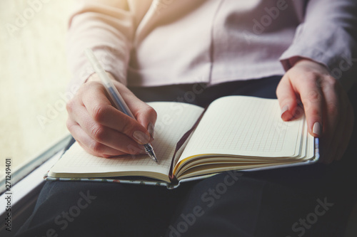 young woman sitting next to the window writing notes on notebook with pen