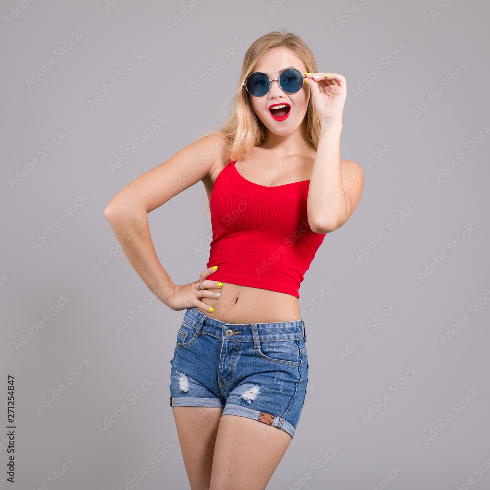 Surprised young woman with sunglasses in denim shorts and red top on light grey background. Summer Women's fashion.
