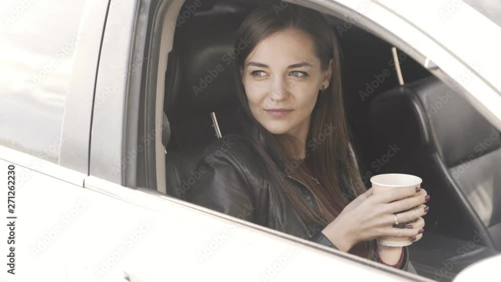 Young woman sitting in car and holding coffee in her hands, closes car window Young woman sitting in car and holding coffee in her hands, closes car window
