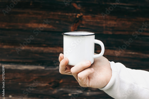 Close up of man travelers hand holding metal mug with hot drink. Outdoor tea, coffee relax time. Mockup of white enamel cup. Adventure, travel, tourism and camping concept. Blurred wooden background. photo