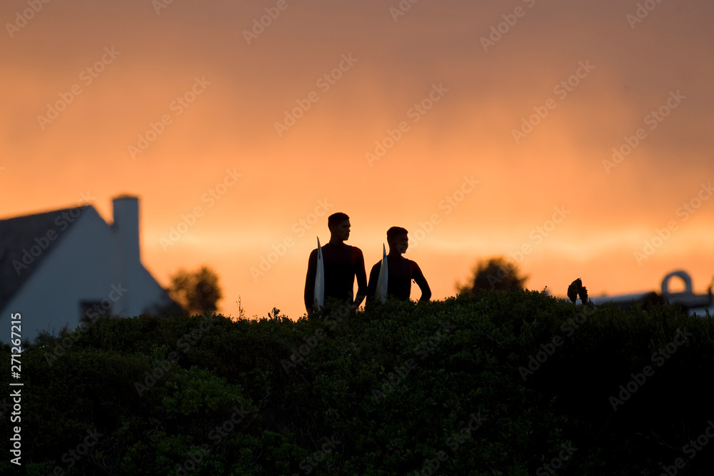 Two surfers stand watching the waves at sunset