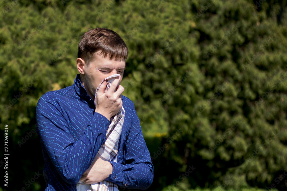 Closeup portrait of a young man with allergies. A man sneezes into a scarf on the background of green trees. Closeup portrait of a young man with allergies. A man sneezes into a scarf on the background of green trees.