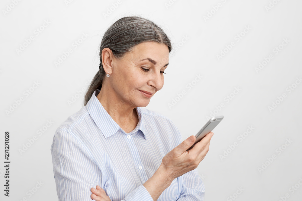 People, age and gadgets concept. Joyful positive mature businesswoman with gray hair gathered in ponytail scrolling social media newsfeed with curious smile, using mobile phone during break People, age and gadgets concept. Joyful positive mature businesswoman with gray hair gathered in ponytail scrolling social media newsfeed with curious smile, using mobile phone during break