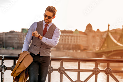 An elegant man standing next to a river and checking the time