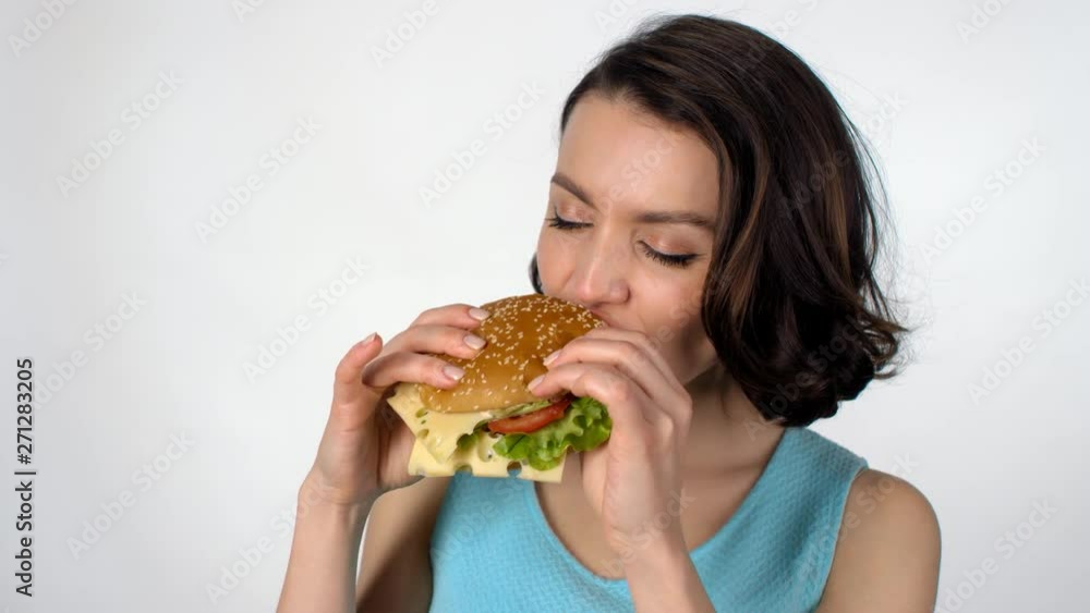 Portrait of beautiful young woman standing isolated on white background and eating delicious burger while looking at camera Portrait of beautiful young woman standing isolated on white background and eating delicious burger while looking at camera