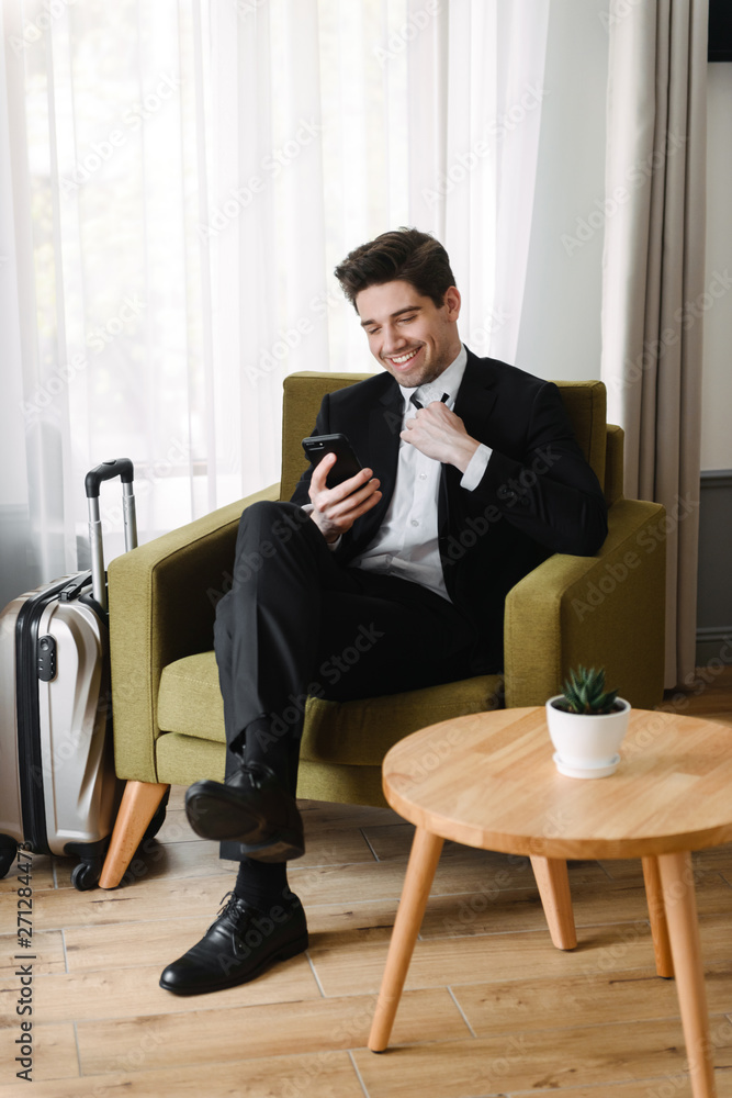 Photo of happy calming businessman making video call on cellphone while sitting on armchair in hotel apartment Photo of happy calming businessman making video call on cellphone while sitting on armchair in hotel apartment