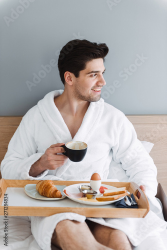 Photo of unshaven caucasian man having breakfast while sitting on bed in hotel apartment