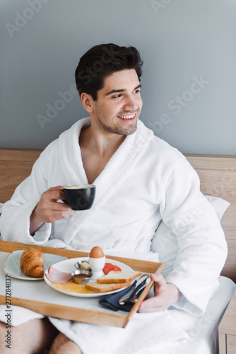 Photo of happy caucasian man wearing white bathrobe having breakfast in hotel apartment