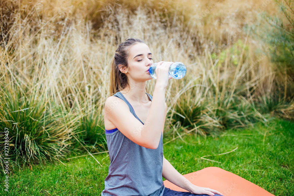 Young woman sitting on yoga mat and drinking water from bottle in summer day Young woman sitting on yoga mat and drinking water from bottle in summer day