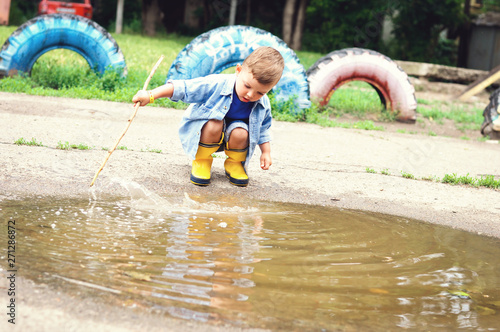 Fototapeta Naklejka Na Ścianę i Meble -  A three-year-old boy in yellow rubber boots and a denim blue shirt plays with a stick in a puddle
