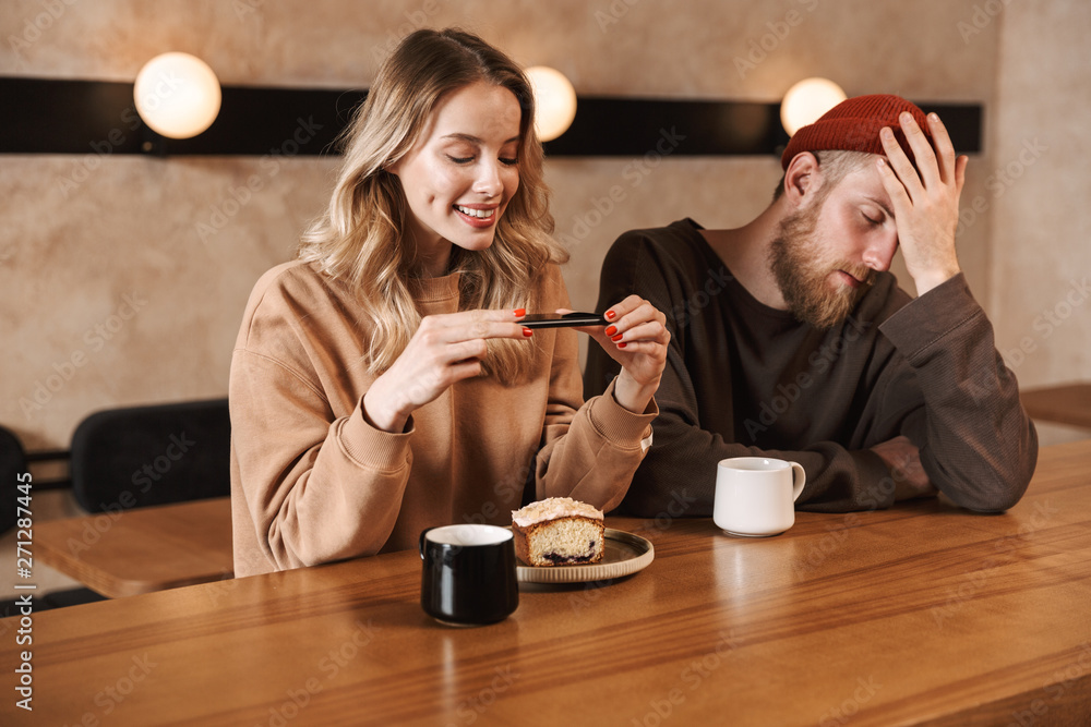 Happy young woman take photo of breakfast nearher displeased man in cafe drinking coffee. Happy young woman take photo of breakfast nearher displeased man in cafe drinking coffee.