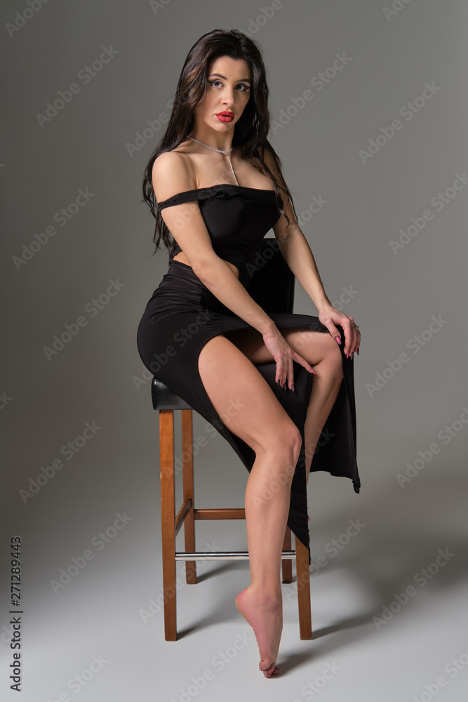 Fototapeta premium Young attractive brunette sexy woman sits on chair in erotic black cocktail dress. Gray background