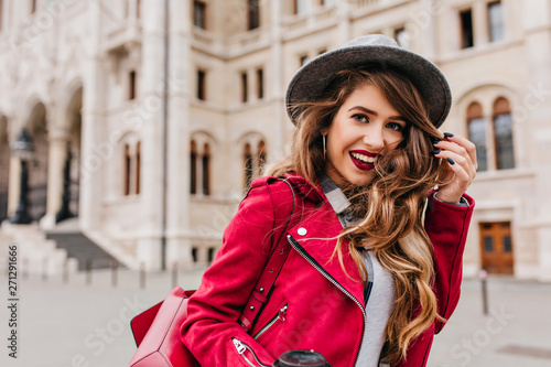 Amazing white female model playing with her long wavy hair while walking past old house. Outdoor shot of refined girl in red casual jacket expressing positive emotions in weekend in Europe.