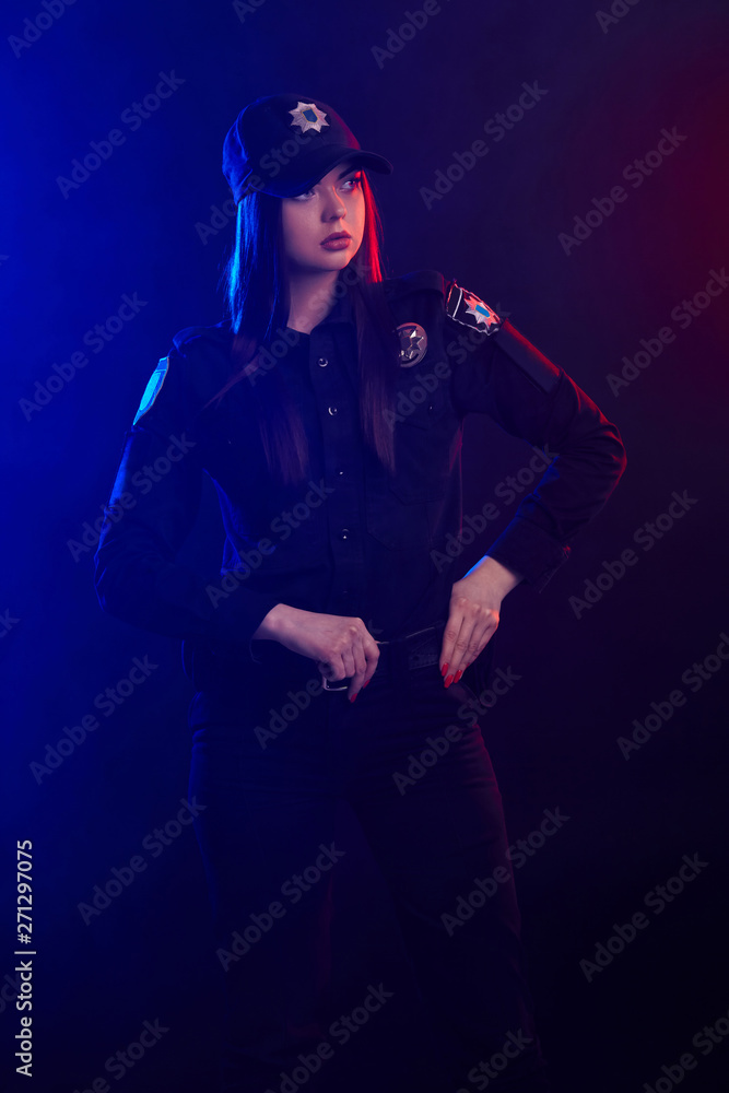 Serious female police officer is posing for the camera against a black background with red and blue backlighting. Serious female police officer is posing for the camera against a black background with red and blue backlighting.