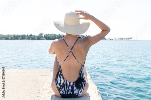 Young woman sitting on beach holding beach hat enjoying summer holidays looking at the sea. Beautiful back side of model in bikini sitting down
