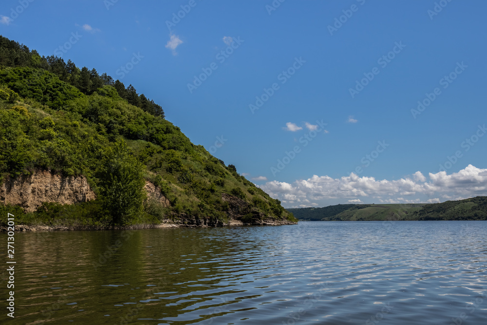 The high and steep bank of the Dniester, covered with green grass, shrubs and forest. Clear sky and clouds on the horizon. Summer landscape. Ukraine.