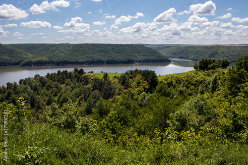 The narrow strip of water of the Dniester River is clamped between the high hills of the shores covered with green forest. The sky is covered with white clouds. Summer landscape. Ukraine.