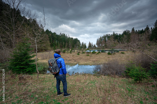 A man standing in front of a forest and a lake during a cloudy day in mountain