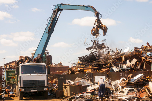 A grapple truck and a man in an orange helmet work on a hot sunny day at a large scrap yard.