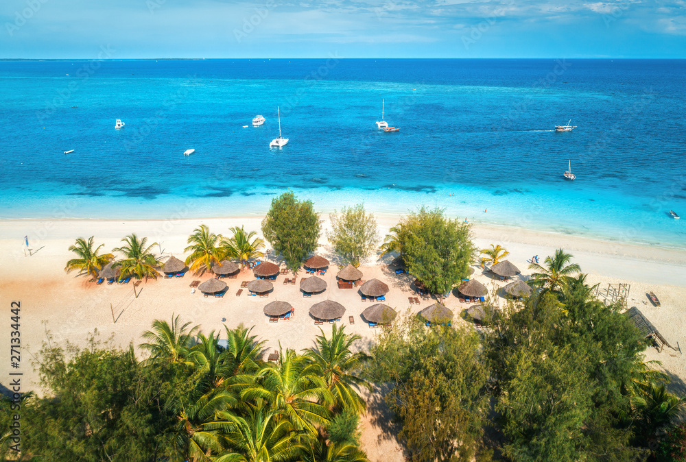 Aerial view of umbrellas, palms on the sandy beach of Indian Ocean at sunny day. Summer holiday in Zanzibar, Africa. Tropical landscape with palm trees, parasols, boats, yachts, blue water. Top view