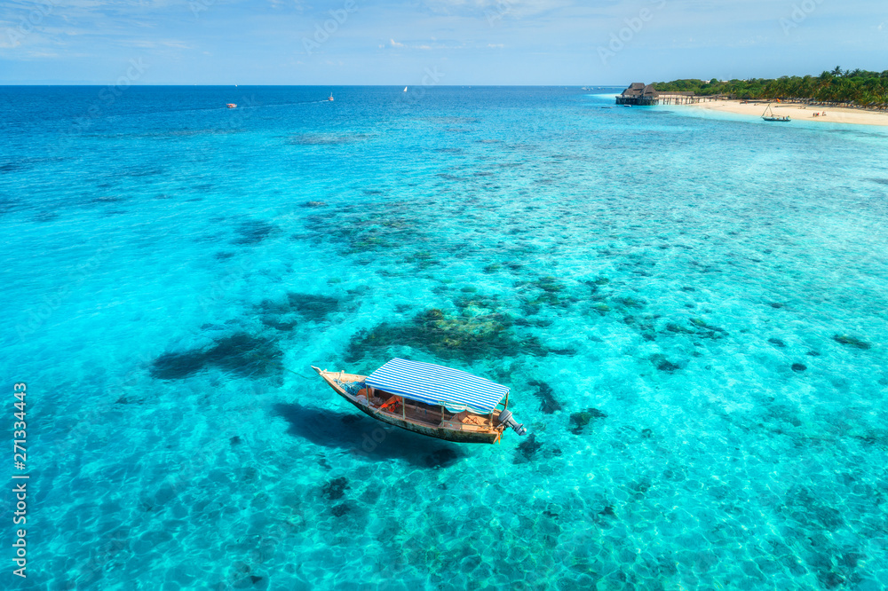 Fototapeta premium Aerial view of the fishing boat in clear blue water at sunny day in summer. Top view from the air of boat, sandy beach. Indian ocean in Zanzibar, Africa. Colorful Landscape with motorboat, clear sea